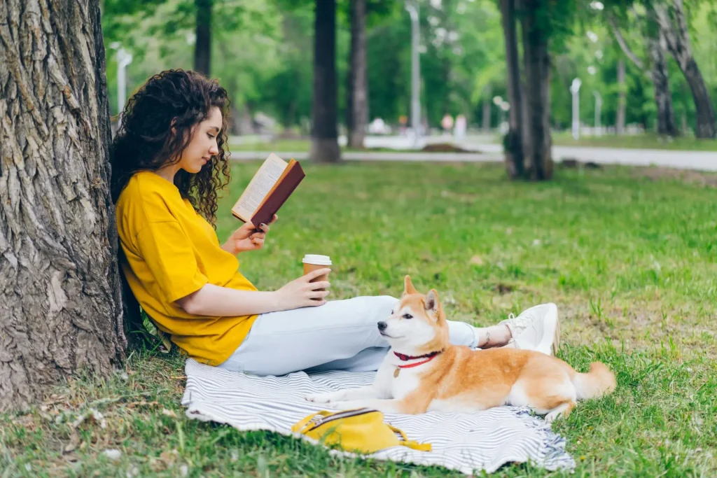 Woman sitting in a blanket reading a book in a park accompanied by her dog